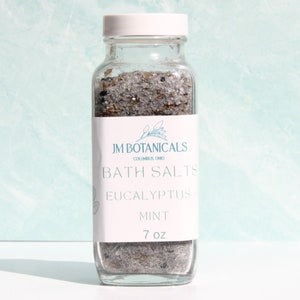 Puede incluir: Un frasco de vidrio transparente lleno de sales de baño. La etiqueta dice "JM Botanicals, Columbus, Ohio, Bath Salts, Eucalyptus Mint, 7 oz."