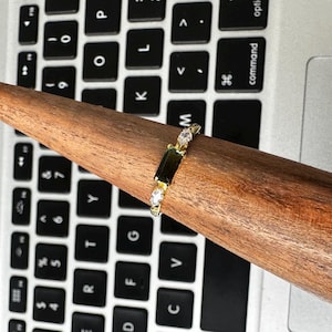 Puede incluir: Un anillo de oro con una gema verde rectangular y dos gemas blancas m&aacute;s peque&ntilde;as. El anillo se muestra en un soporte de madera para anillos, sobre un teclado de port&aacute;til.
