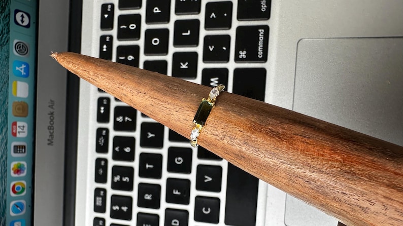 Puede incluir: Un anillo de oro con una gema verde rectangular y dos gemas blancas m&aacute;s peque&ntilde;as. El anillo se muestra en un soporte de madera para anillos, sobre un teclado de port&aacute;til.
