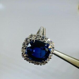 May include: A silver-toned ring featuring a large, cushion-cut deep blue sapphire center stone. The sapphire is surrounded by a halo of small, round, clear white stones. The band is also accented with small, clear white stones along the top shank.