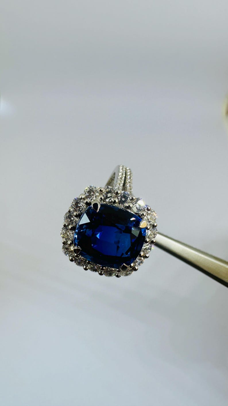 May include: A silver-toned ring featuring a large, cushion-cut deep blue sapphire center stone. The sapphire is surrounded by a halo of small, round, clear white stones. The band is also accented with small, clear white stones along the top shank.