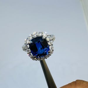May include: A silver-toned ring featuring a large, cushion-cut deep blue gemstone at the center, surrounded by a halo of small, clear, round-cut stones. The band is also adorned with a row of small, clear, round-cut stones along its visible sides.