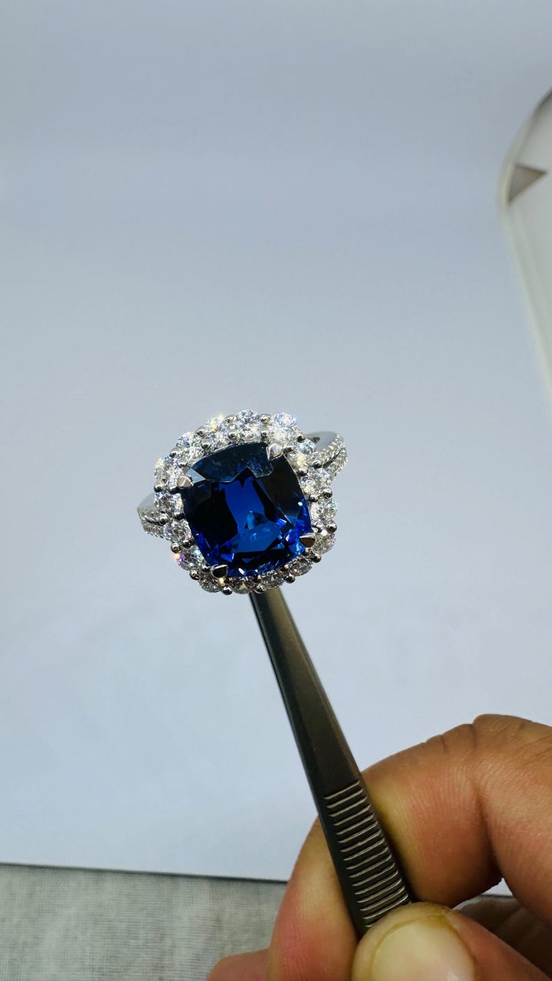 May include: A silver-toned ring featuring a large, cushion-cut deep blue gemstone at the center, surrounded by a halo of small, clear, round-cut stones. The band is also adorned with a row of small, clear, round-cut stones along its visible sides.