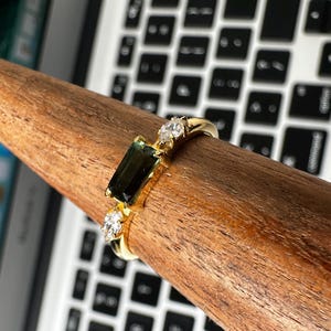 Puede incluir: Un anillo de oro con una piedra rectangular verde oscuro y dos piedras claras m&aacute;s peque&ntilde;as. El anillo se muestra sobre un objeto de madera, con un teclado de port&aacute;til al fondo. El anillo es una joya.