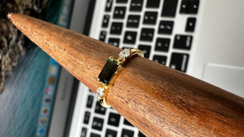 Puede incluir: Un anillo de oro con una piedra rectangular verde oscuro y dos piedras claras m&aacute;s peque&ntilde;as. El anillo se muestra sobre un objeto de madera, con un teclado de port&aacute;til al fondo. El anillo es una joya.