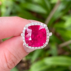 May include: A cushion-cut ruby ring with a halo of small diamonds. The ruby is a deep red color, and the diamonds sparkle around it. The ring is set in a white metal, and the background is blurred green foliage.