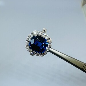 May include: A silver-toned ring featuring a large, cushion-cut deep blue gemstone at the center, surrounded by a halo of small, clear, round-cut white stones. The ring band is thin and metallic, held by metal tweezers for display purposes.