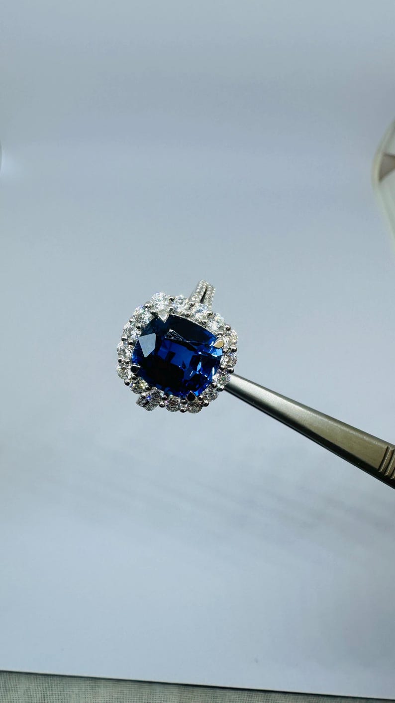 May include: A silver-toned ring featuring a large, cushion-cut deep blue gemstone at the center, surrounded by a halo of small, clear, round-cut white stones. The ring band is thin and metallic, held by metal tweezers for display purposes.