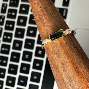 Puede incluir: Un anillo de oro con una piedra preciosa rectangular de color verde oscuro y peque&ntilde;as piedras claras. El anillo se muestra en un soporte de madera, con un teclado de port&aacute;til en el fondo.
