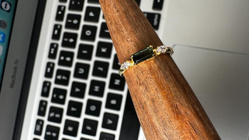 Puede incluir: Un anillo de oro con una piedra preciosa rectangular de color verde oscuro y peque&ntilde;as piedras claras. El anillo se muestra en un soporte de madera, con un teclado de port&aacute;til en el fondo.