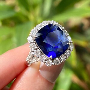 May include: A ring featuring a large, cushion-cut deep blue sapphire center stone surrounded by a halo of round brilliant-cut white diamonds. The band is crafted from a silver-toned metal and includes additional diamonds along the shank.