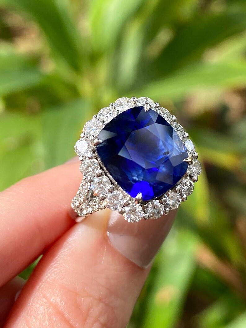 May include: A ring featuring a large, cushion-cut deep blue sapphire center stone surrounded by a halo of round brilliant-cut white diamonds. The band is crafted from a silver-toned metal and includes additional diamonds along the shank.