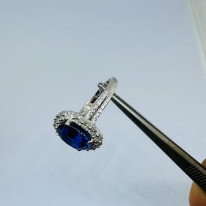 May include: A silver-toned ring featuring a large, oval-cut deep blue center stone surrounded by a halo of small, clear, sparkling stones. The band is split and embellished with additional clear stones along the sides of the setting.