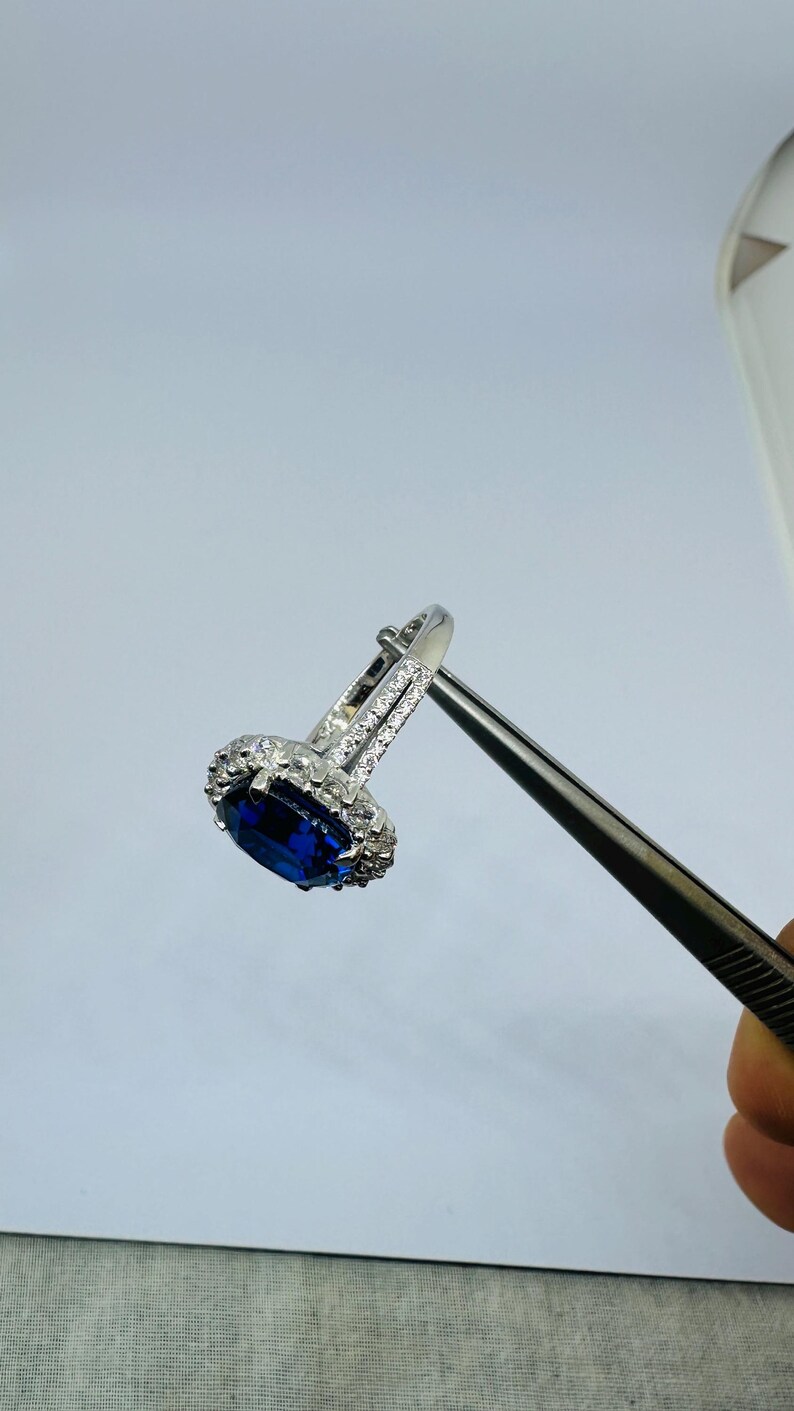 May include: A silver-toned ring featuring a large, oval-cut deep blue center stone surrounded by a halo of small, clear, sparkling stones. The band is split and embellished with additional clear stones along the sides of the setting.