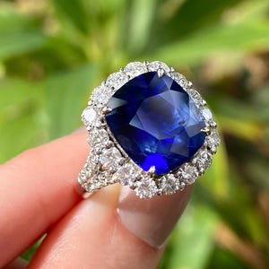 May include: A ring featuring a large, cushion-cut deep blue sapphire center stone surrounded by a halo of round brilliant-cut white diamonds. The band is crafted from a silver-toned metal and includes additional diamonds along the shank.