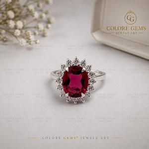 May include: A silver ring featuring a large, oval-cut, deep red gemstone at the centre, surrounded by a halo of small, round, clear white stones. The band is polished silver. The text COLORE GEMS JEWELS ART is visible in the upper right corner.