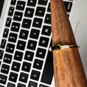 Puede incluir: Un anillo de oro con una piedra rectangular verde oscuro y dos piedras blancas m&aacute;s peque&ntilde;as. El anillo se exhibe en un soporte de madera, con un teclado de port&aacute;til en el fondo.
