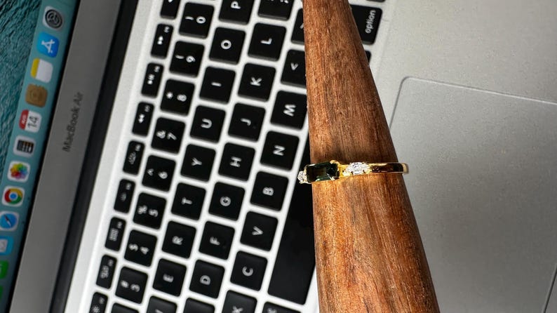 Puede incluir: Un anillo de oro con una piedra rectangular verde oscuro y dos piedras blancas m&aacute;s peque&ntilde;as. El anillo se exhibe en un soporte de madera, con un teclado de port&aacute;til en el fondo.