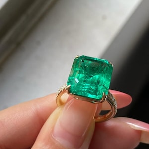 May include: A gold ring with a large, rectangular emerald gemstone. The emerald is a vivid green and set in a gold band with small accent stones. The ring is a striking piece of jewellery.