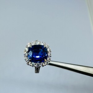 May include: A silver-toned ring featuring a large, cushion-cut deep blue gemstone at the center, surrounded by a halo of small, clear, round-cut white stones. The band is also accented with small white stones along its visible upper shank.