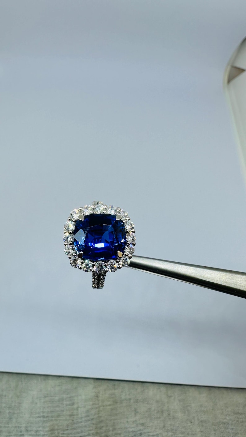 May include: A silver-toned ring featuring a large, cushion-cut deep blue gemstone at the center, surrounded by a halo of small, clear, round-cut white stones. The band is also accented with small white stones along its visible upper shank.