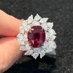 May include: A ring featuring a large, oval-cut deep red ruby at the center, surrounded by a double halo of numerous marquise-cut, clear white diamonds set in a metallic band. The design creates a floral-like arrangement of sparkling stones.