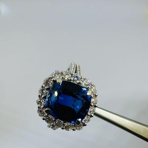 May include: A silver-toned ring featuring a large, cushion-cut deep blue gemstone at the center. The central stone is surrounded by a halo of small, clear, round-cut stones. The band is thin and also accented with small, clear, round-cut stones.