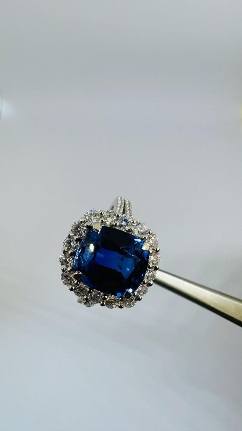 May include: A silver-toned ring featuring a large, cushion-cut deep blue gemstone at the center. The central stone is surrounded by a halo of small, clear, round-cut stones. The band is thin and also accented with small, clear, round-cut stones.