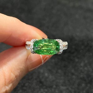 May include: A close-up of a ring featuring a large, rectangular, vibrant green gemstone. The gemstone is surrounded by smaller, clear stones set in a silver or white gold band. The ring is held up against a dark background.