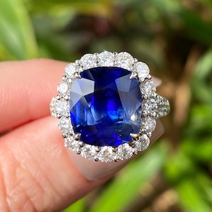 May include: A cushion-cut deep blue sapphire ring featuring a halo of round brilliant-cut diamonds set in a silver-toned metal band. The central gemstone is large and faceted, surrounded by a scalloped border of smaller sparkling white diamonds.