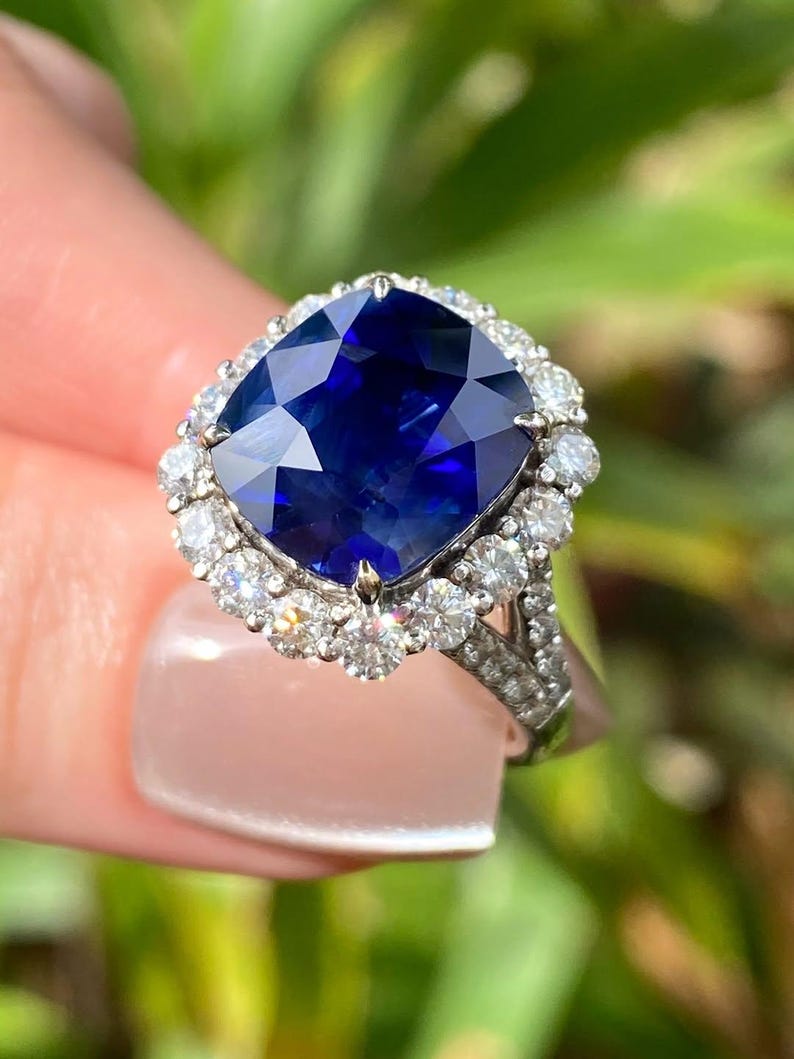 May include: A cushion-cut deep blue sapphire ring set in a silver-toned metal band. The central sapphire is surrounded by a halo of small, round, clear white diamonds, with additional small diamonds embedded along the split shank of the ring band.