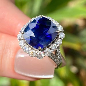 May include: A cushion-cut deep blue sapphire ring set in a silver-toned metal band. The central sapphire is surrounded by a halo of small, round, clear white diamonds, with additional small diamonds embedded along the split shank of the ring band.