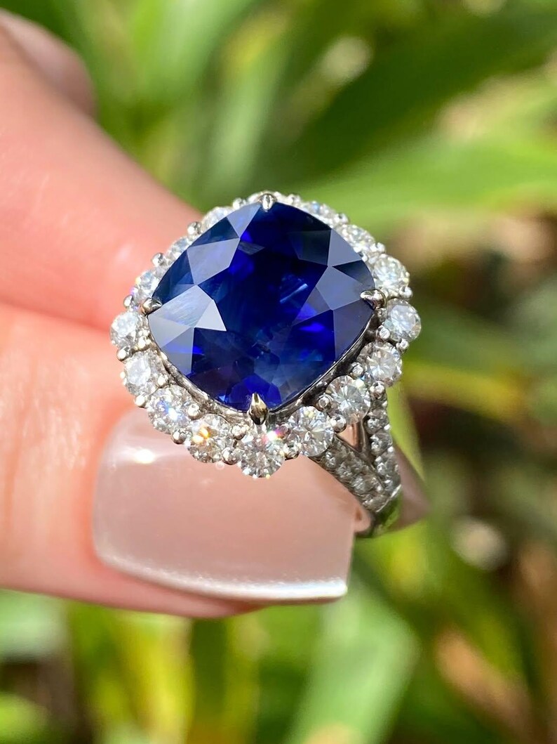 May include: A cushion-cut deep blue sapphire ring set in a silver-toned metal band. The central sapphire is surrounded by a halo of small, round, clear white diamonds, with additional small diamonds embedded along the split shank of the ring band.