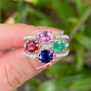 May include: Five gemstone rings stacked together. Each ring features a different coloured gemstone: pink, red, green, and blue. The gemstones are set in silver bands with small, clear stones.