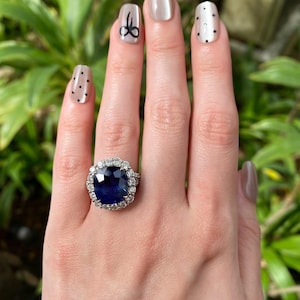 May include: A ring featuring a large, cushion-cut deep blue sapphire center stone surrounded by a halo of round brilliant-cut white diamonds. The setting is crafted from a polished silver-toned metal, showcasing a classic cluster design.