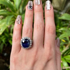 May include: A ring featuring a large, cushion-cut deep blue sapphire center stone surrounded by a halo of round brilliant-cut white diamonds. The setting is crafted from a polished silver-toned metal, showcasing a classic cluster design.