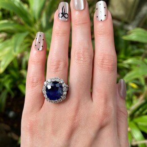 May include: A ring featuring a large, cushion-cut deep blue sapphire center stone surrounded by a halo of round brilliant-cut white diamonds. The setting is crafted from a polished silver-toned metal, showcasing a classic cluster design.