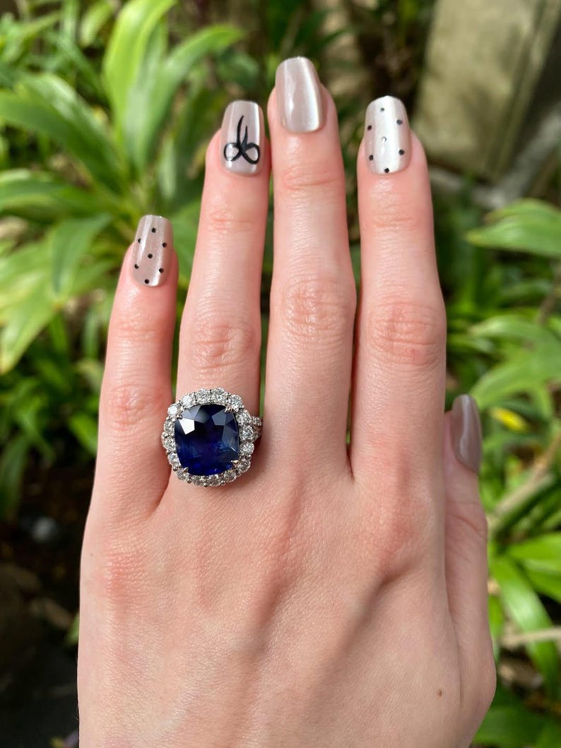 May include: A ring featuring a large, cushion-cut deep blue sapphire center stone surrounded by a halo of round brilliant-cut white diamonds. The setting is crafted from a polished silver-toned metal, showcasing a classic cluster design.