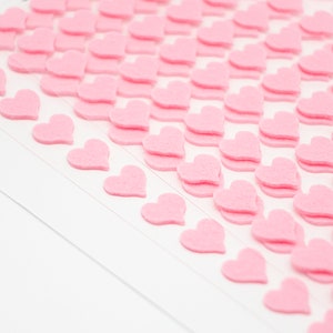 K&ouml;nnte beinhalten: Nahaufnahme von kleinen, rosa Filz-Herz-Aufklebern, die in Reihen auf einer wei&szlig;en Oberfl&auml;che angeordnet sind. Die Aufkleber sind geschichtet und erzeugen einen 3D-Effekt. Eine kleine Illustration eines Charakters, der ein Herz h&auml;lt, ist sichtbar.