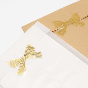 Pu&ograve; includere: Una busta in carta bianca traslucida e una busta in carta kraft marrone, ciascuna con un adesivo in lamina d'oro a forma di fiocco. Gli adesivi a fiocco dorato presentano un motivo a righe lineari e sono centrati sulla patta di ogni busta.