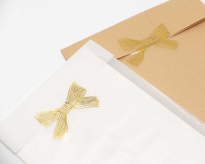 Pu&ograve; includere: Una busta in carta bianca traslucida e una busta in carta kraft marrone, ciascuna con un adesivo in lamina d'oro a forma di fiocco. Gli adesivi a fiocco dorato presentano un motivo a righe lineari e sono centrati sulla patta di ogni busta.