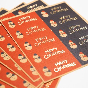 Può includere: Una collezione di fogli di adesivi rettangolari rossi e neri, ciascuno con un motivo ripetitivo di un piccolo pupazzo di neve con cappello e il testo Merry Christmas stampato in un carattere stilizzato in griglia.