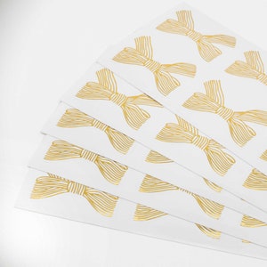 Pu&ograve; includere: Una pila di fogli di carta rettangolari bianchi con un motivo ripetuto di fiocchi disegnati a linee dorate. Ogni fiocco &egrave; realizzato con sottili linee orizzontali parallele che creano un aspetto metallico e strutturato sullo sfondo bianco.