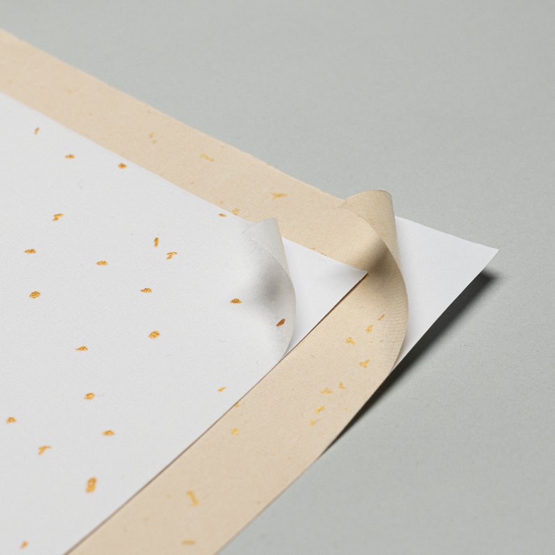 May include: Two sheets of paper with gold flecks. The top sheet is white and has a corner folded back to reveal the beige sheet underneath.