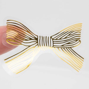 Pu&ograve; includere: Un adesivo trasparente con un design a fiocco in oro metallizzato. Il fiocco &egrave; composto da sottili linee dorate parallele che formano la sagoma di un nastro annodato, con un nodo centrale e due asole su ciascun lato su supporto trasparente.