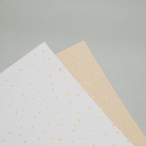 May include: Two sheets of paper with a speckled pattern. One sheet is white with gold flecks, and the other is beige with gold flecks.