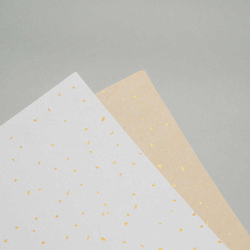 May include: Two sheets of paper with a speckled pattern. One sheet is white with gold flecks, and the other is beige with gold flecks.
