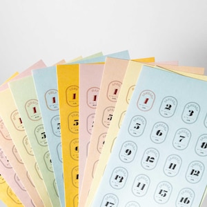 May include: A set of colorful paper sheets with stickers featuring the months of the year and numbers 1 through 17. The stickers are in a vintage style with a gold border and black text.