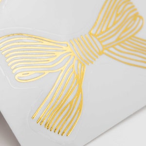 Pu&ograve; includere: Un adesivo in lamina d'oro con un design a fiocco stilizzato composto da sottili linee parallele. Il fiocco &egrave; stampato su un supporto adesivo trasparente, mostrando una finitura metallica che riflette la luce sul suo intricato motivo lineare.