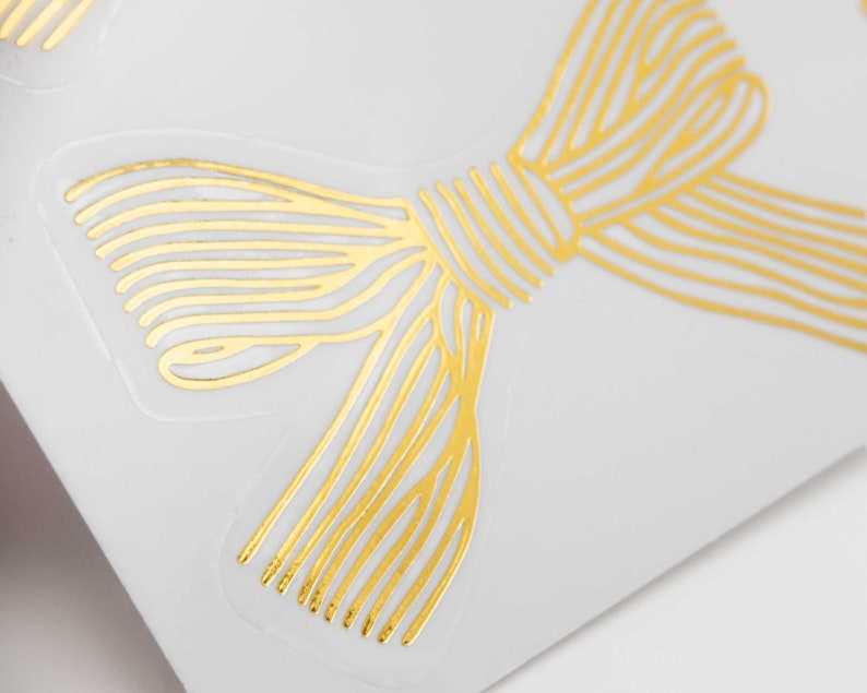 Pu&ograve; includere: Un adesivo in lamina d'oro con un design a fiocco stilizzato composto da sottili linee parallele. Il fiocco &egrave; stampato su un supporto adesivo trasparente, mostrando una finitura metallica che riflette la luce sul suo intricato motivo lineare.